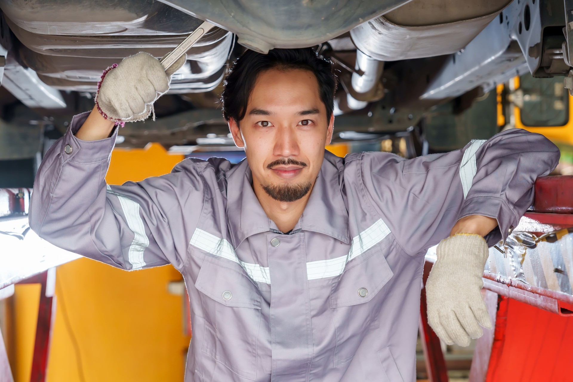 Mecánico sonriendo bajo el elevador de coches con herramientas en la mano, enfatizando la confianza, el profesionalismo y la dedicación a la calidad en la reparación de automóviles en un taller de confianza para pequeñas empresas.